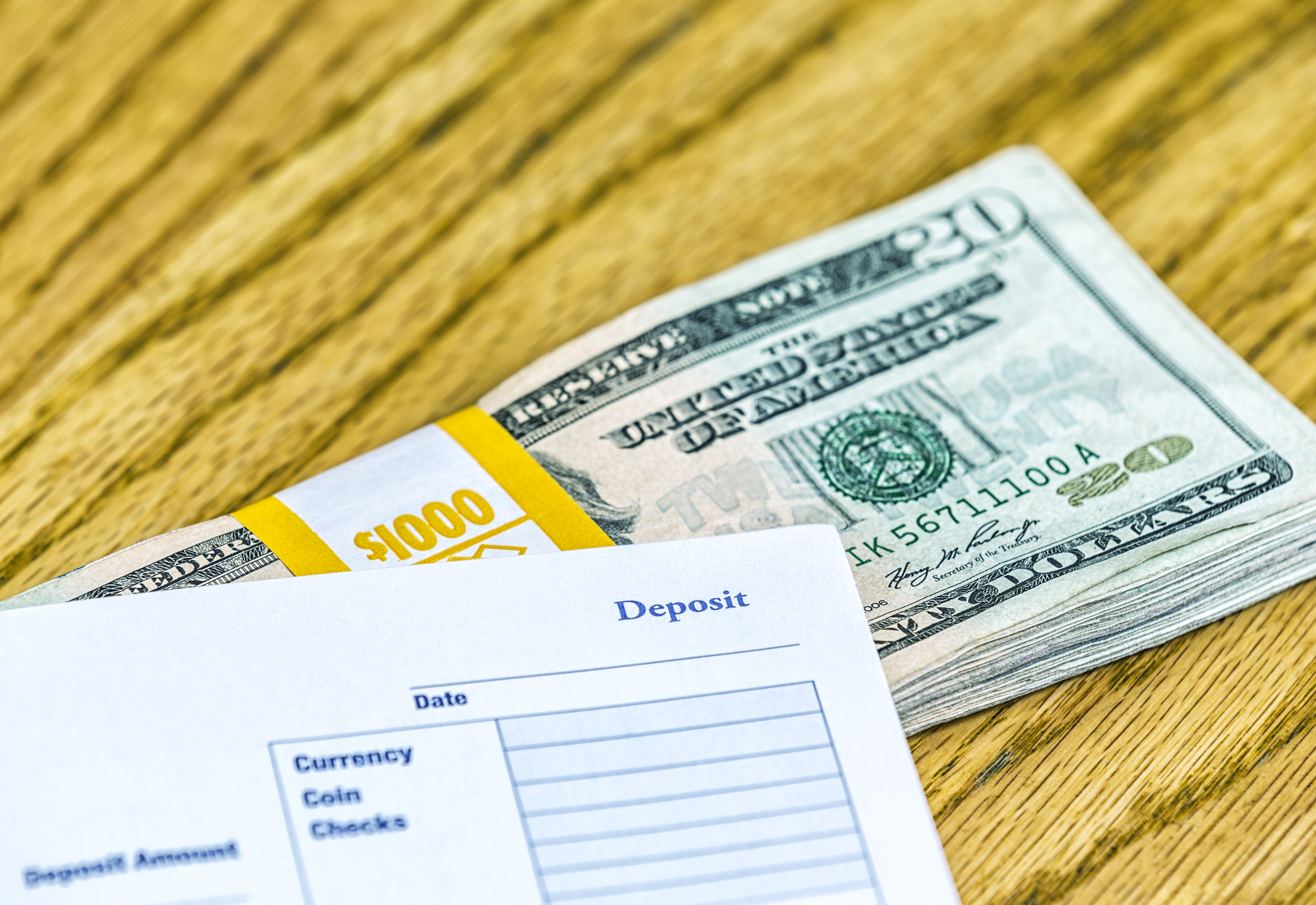 One thousand dollars in a wrapped stack of USA twenty dollar bills and a banking account deposit slip on a wooden table top.